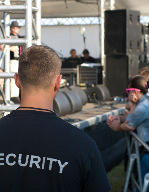 security personnel overseeing a live event with a crowd in the background and sound equipment visible